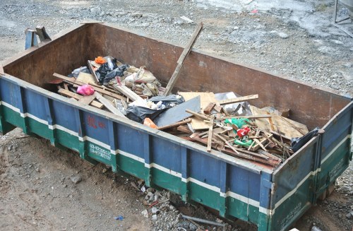 A large, rectangular metal skip container situated outdoors on a gravel and dirt surface, filled with assorted construction and renovation debris including wooden planks, broken panels, plastic wrappers, and small fragmented materials. The skip's sides are painted in a blue and green colour scheme, with visible signs of wear and rust around the edges, indicating it is used for waste removal. In the background, a rough, unpaved ground area is visible, suggesting a site undergoing building work or office clearance. The debris appears to be a mix of wooden and plastic waste, aligned with typical rubbish removed during estate or office clearances in the Downham area, which is part of London. The setting looks utilitarian, emphasizing the importance of professional rubbish removal services like those provided by Office Clearance Downham to clear construction debris efficiently and safely from commercial or residential sites.