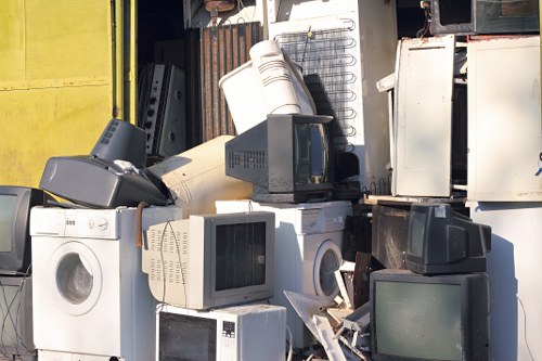 A pile of discarded household appliances and electronic waste, including several computer monitors with dark screens and white casings, a microwave oven, a washing machine with a white metal exterior, and a plastic bucket, all stacked haphazardly outdoors on a driveway or yard area. The background features a partially visible metal structure and a yellow wall, indicating an industrial or waste collection site in the vicinity of Downham, with natural daylight illuminating the scene. The arrangement of the items suggests they are awaiting rubbish removal or disposal, consistent with services offered by Office Clearance Downham for removing unwanted electrical and household items. The textures vary from smooth plastic and metal surfaces to the slightly worn finishes of the appliances, with some items leaning or stacked to maximize space within the confined outdoor environment.