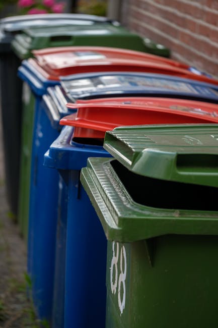 A row of large, rectangular wheelie bins lined up outdoors against a brick wall, with colours including blue, green, and red. The bins are made of durable plastic materials with textured lids, some fe