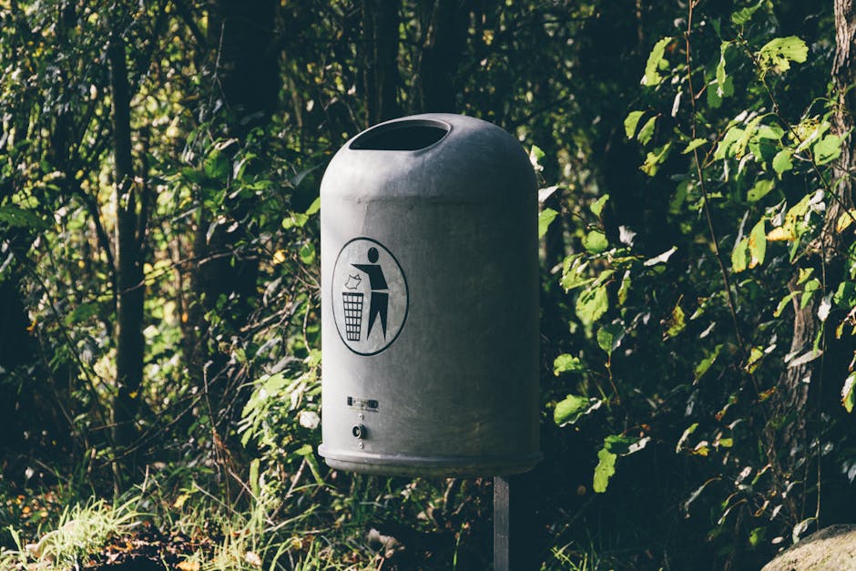 A large, rectangular metal waste bin with a slightly weathered, galvanized finish sits on a paved surface outdoors. The bin has a hinged lid, which appears to be closed, and features a blue and white 